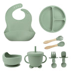 Set of green silicone baby utensils including a bib, plate, cup, and bowls on a white background.