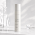 A white cylindrical bottle of THEOBION Barrier Cushion Peptide Cream stands on a light marble surface with soft shadows and blurred glassware in the background.