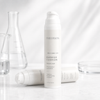A white pump bottle of THEOBION Barrier Cushion Peptide Cream stands upright on a light marble surface, with two additional bottles lying horizontally in the foreground and blurred glassware in the background.